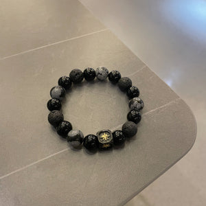 A men's stretch bracelet featuring mixed black lava beads, patterned grey stones, and a central engraved compass cube.